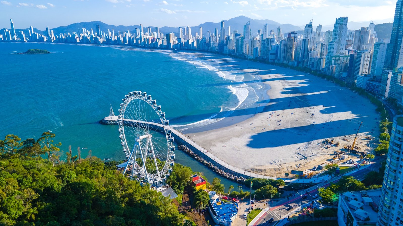 Vista aérea dos arredores da Praia Central. A grande faixa de areia branca é limitada pelo mar azul e por uma fileira de prédios altos. Em uma das extremidades da orla, aparece a Roda Gigante de Balneário Camboriú