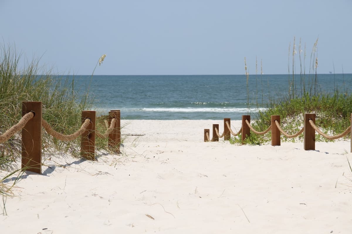 Wooden posts and rope creating a walkway down to the sandy beach with blue waters in the background. Tall green grass to the left and right of the walkway