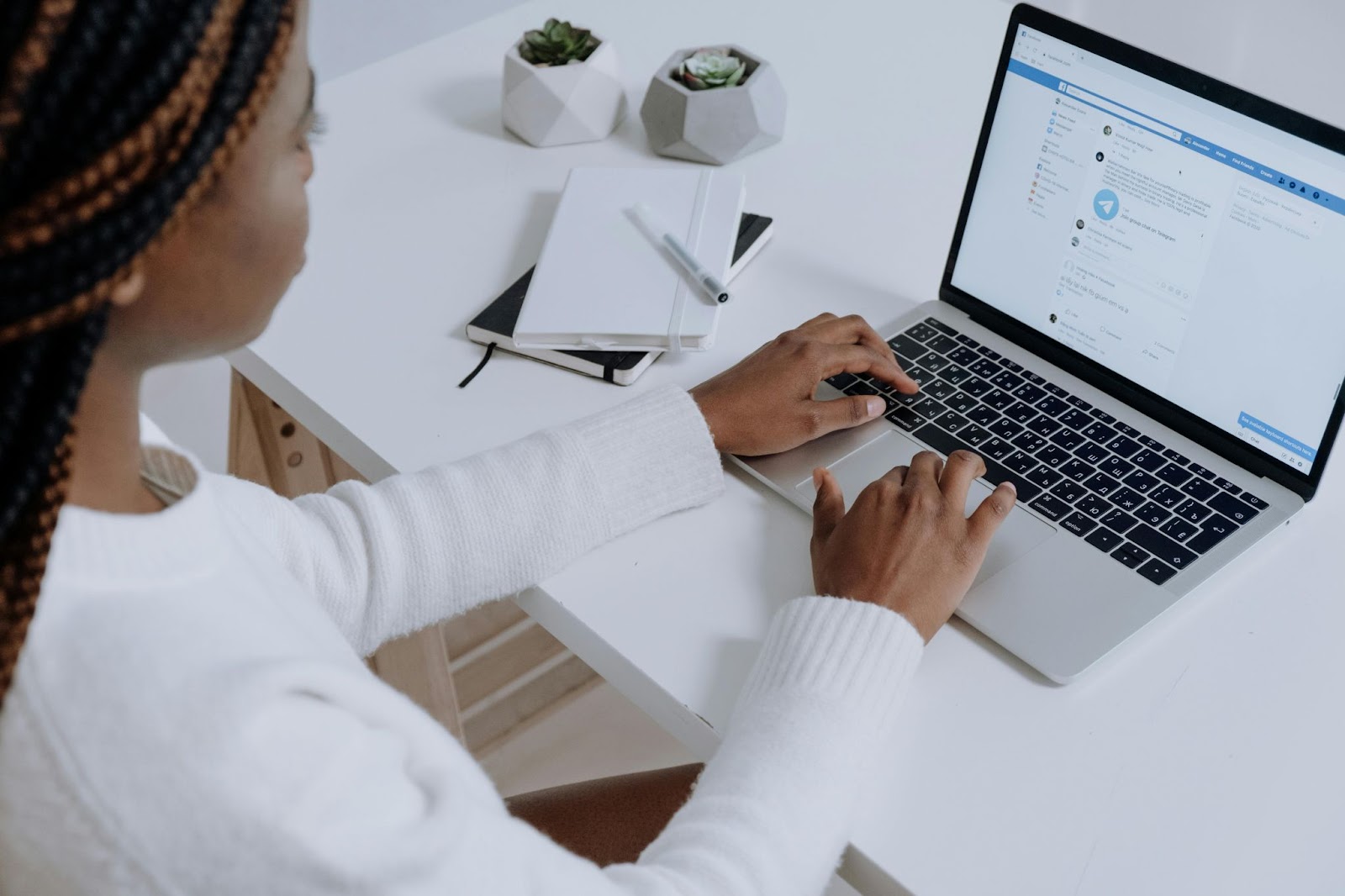 With the laptop screen showing Facebook’s interface, a woman wearing a white sweater scrolls through her feed. Beside the laptop are a couple of succulents in geometric pots and two notebooks stacked on top of each other with a pen on top.
