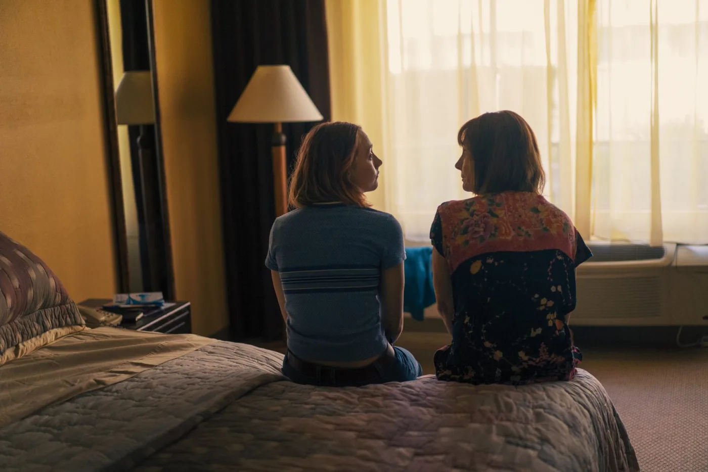 Christine (Saoirse Ronan, a la izquierda) y Marion (Laurie Metcalf, a la derecha) están de espaldas a la cámara mientras conversan en el borde de la cama.