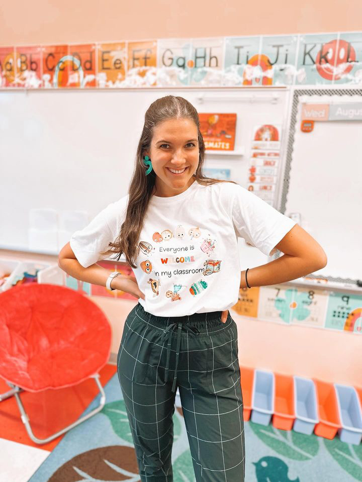 This image shows a smiling teacher in front of a whiteboard in her classroom. You can see fun, bright colors behind her on the whiteboard and the rug. 
