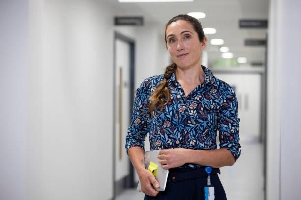 Trustworthy Female Doctor Waist up of a female doctor standing in the hospital corridor. She's dressed in a button-down shirt and has a warm smile on her face and she's holding a digital tablet, she looks directly at the camera, radiating professionalism and a welcoming demeanour. An Insurance Card Holder stock pictures, royalty-free photos & images