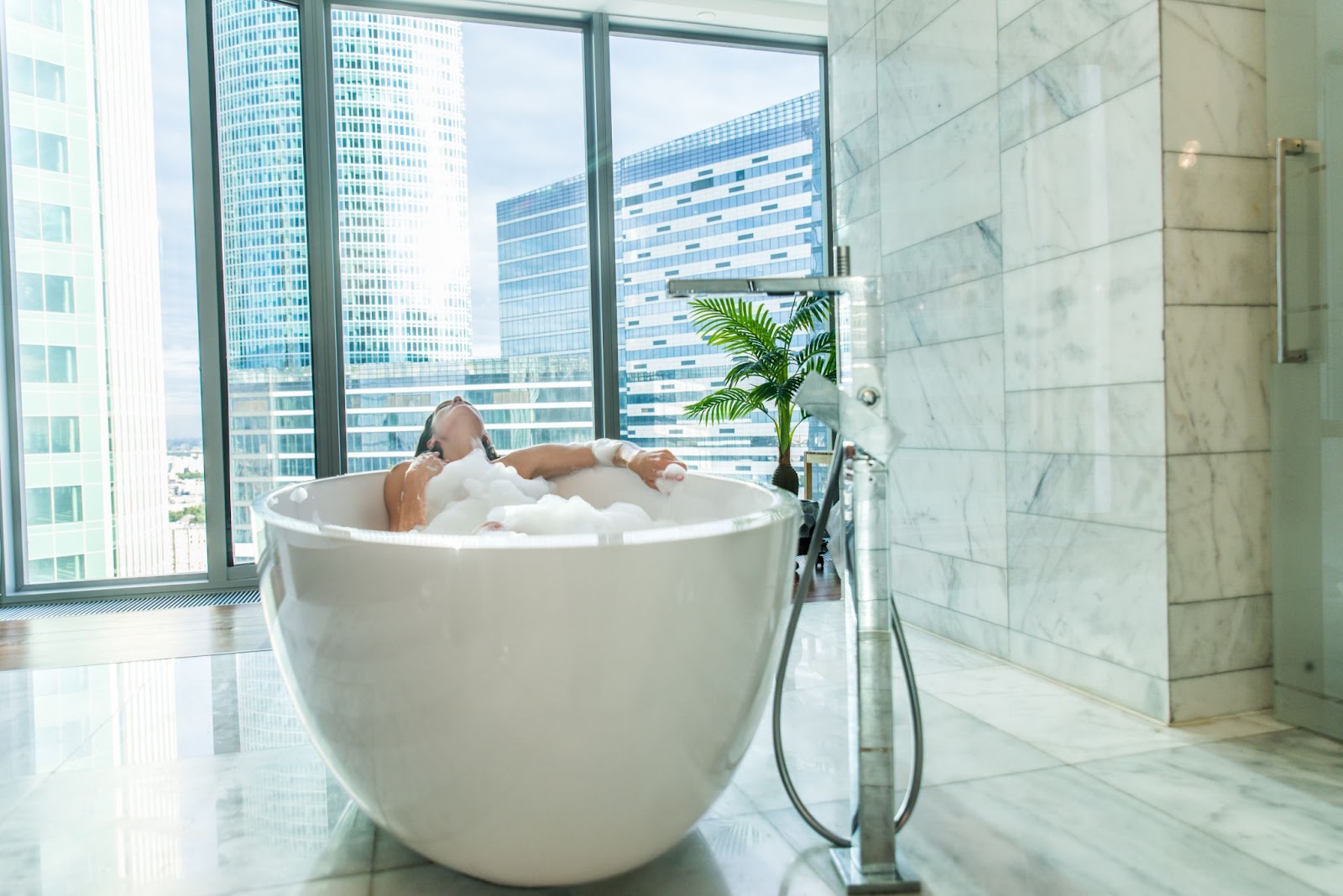 A woman unwinds in a contemporary-style bathtub surrounded by expansive glass walls that offer a stunning view of the urban skyline.