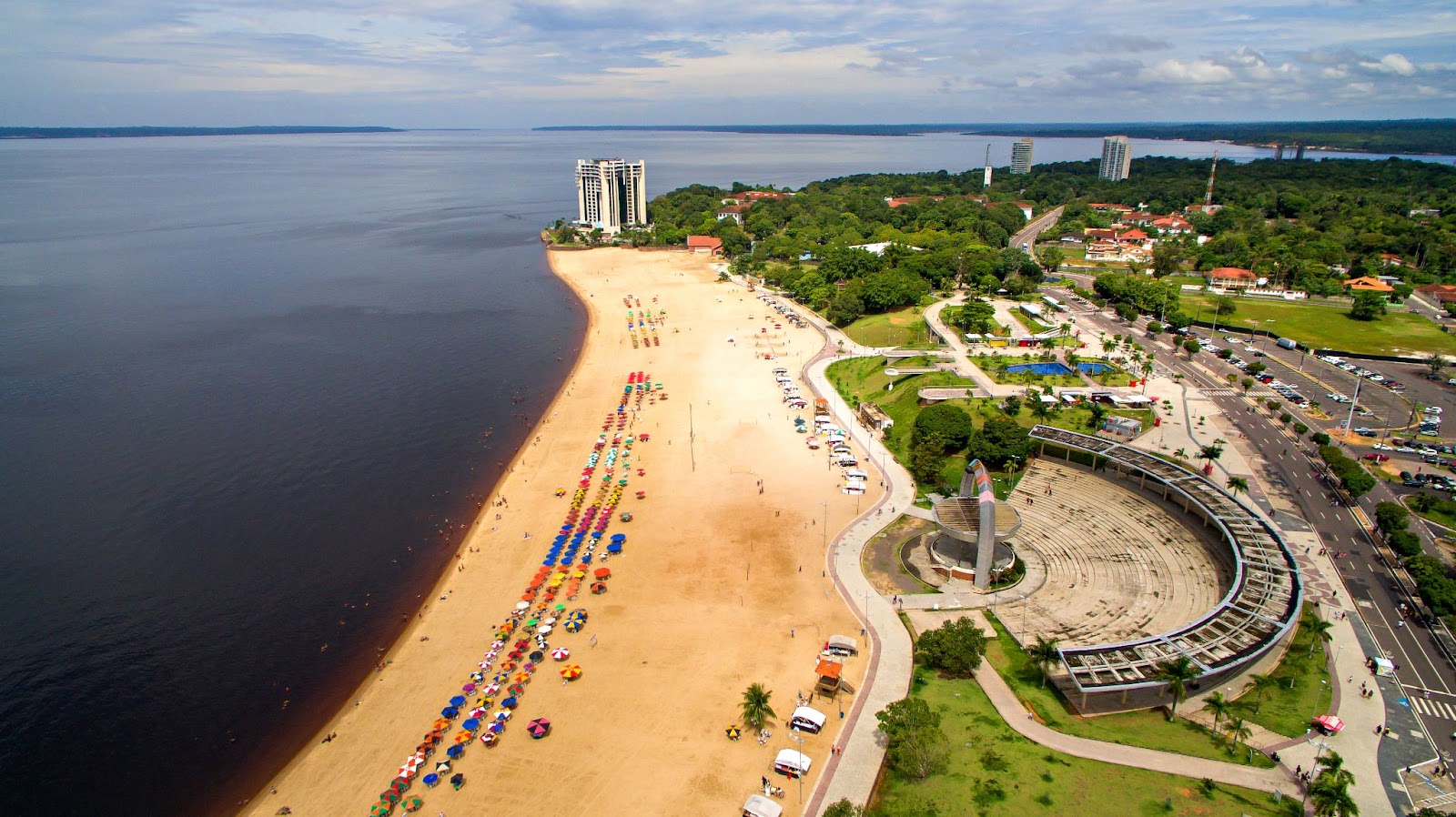 Vista aérea da Praia de Ponta Negra, Manaus. A larga faixa de areia dourada é limitada pelas águas negras do rio e por um longo calçadão onde fica um anfiteatro. Há alguns guarda-sóis e barraquinhas espalhados pela areia