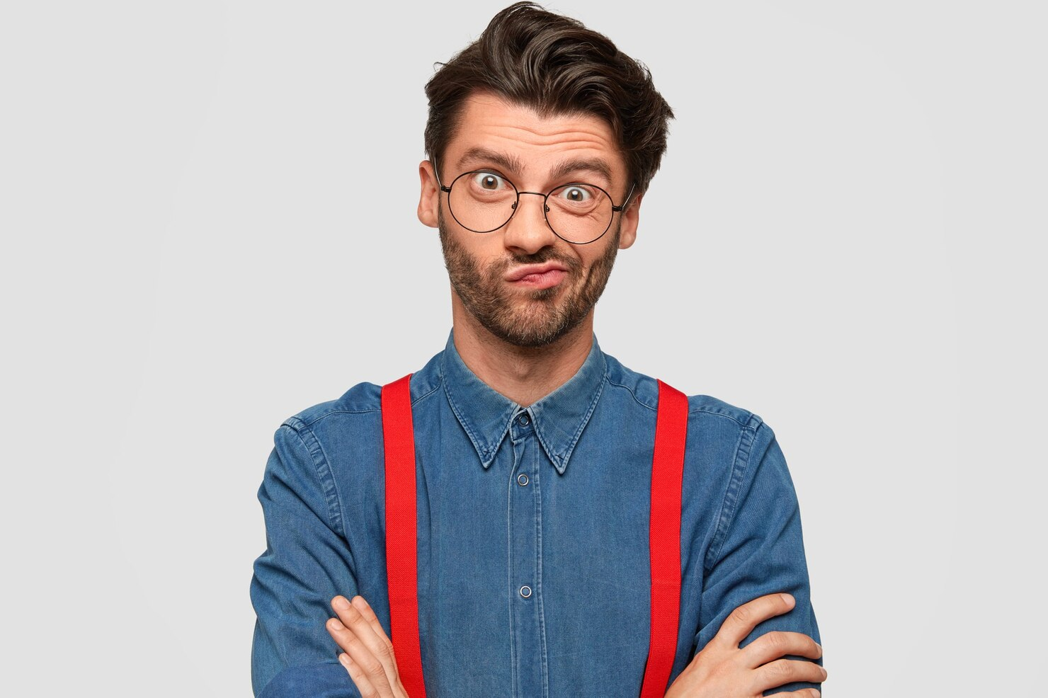 A man in denim shirt and round glasses making dumb facial expressions.