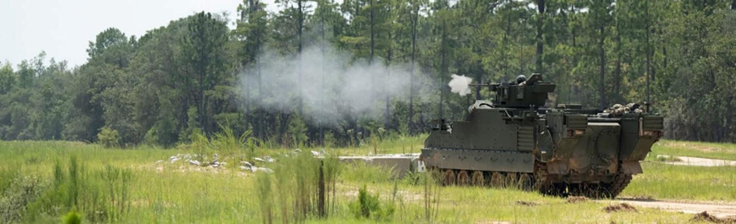 A drab-green Armored Multi-Purpose Vehicle firing rounds while positioned in a grassy field next to a heavily treed forest.