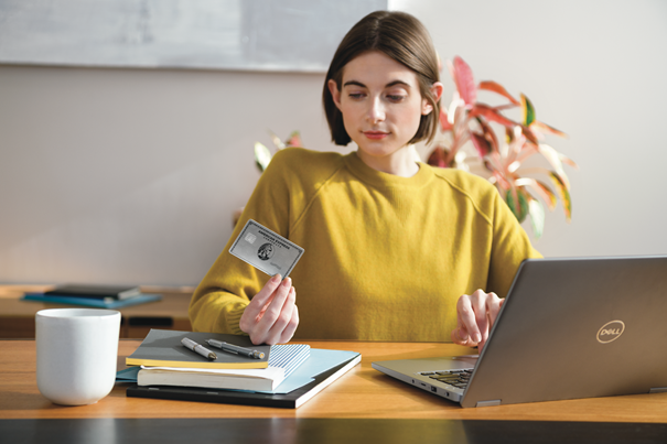 Eine junge Frau mit kinnlangen, dunklen Haaren sitzt an einem Schreibtisch vor ihrem Laptop. Sie hält eine American Express Kreditkarte in ihrer rechten Hand. Auf dem Schreibtisch befinden sich Unterlagen, Stifte und eine Kaffeetasse. 
