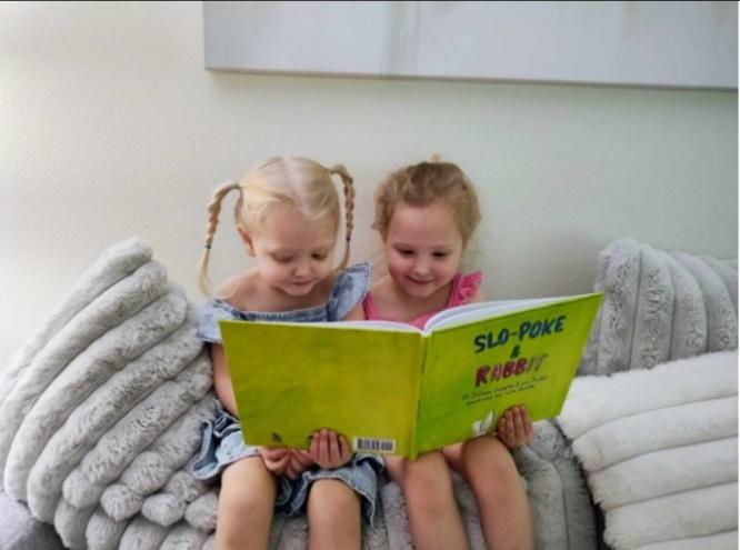 Two girls sitting on a couch reading a book

Description automatically generated