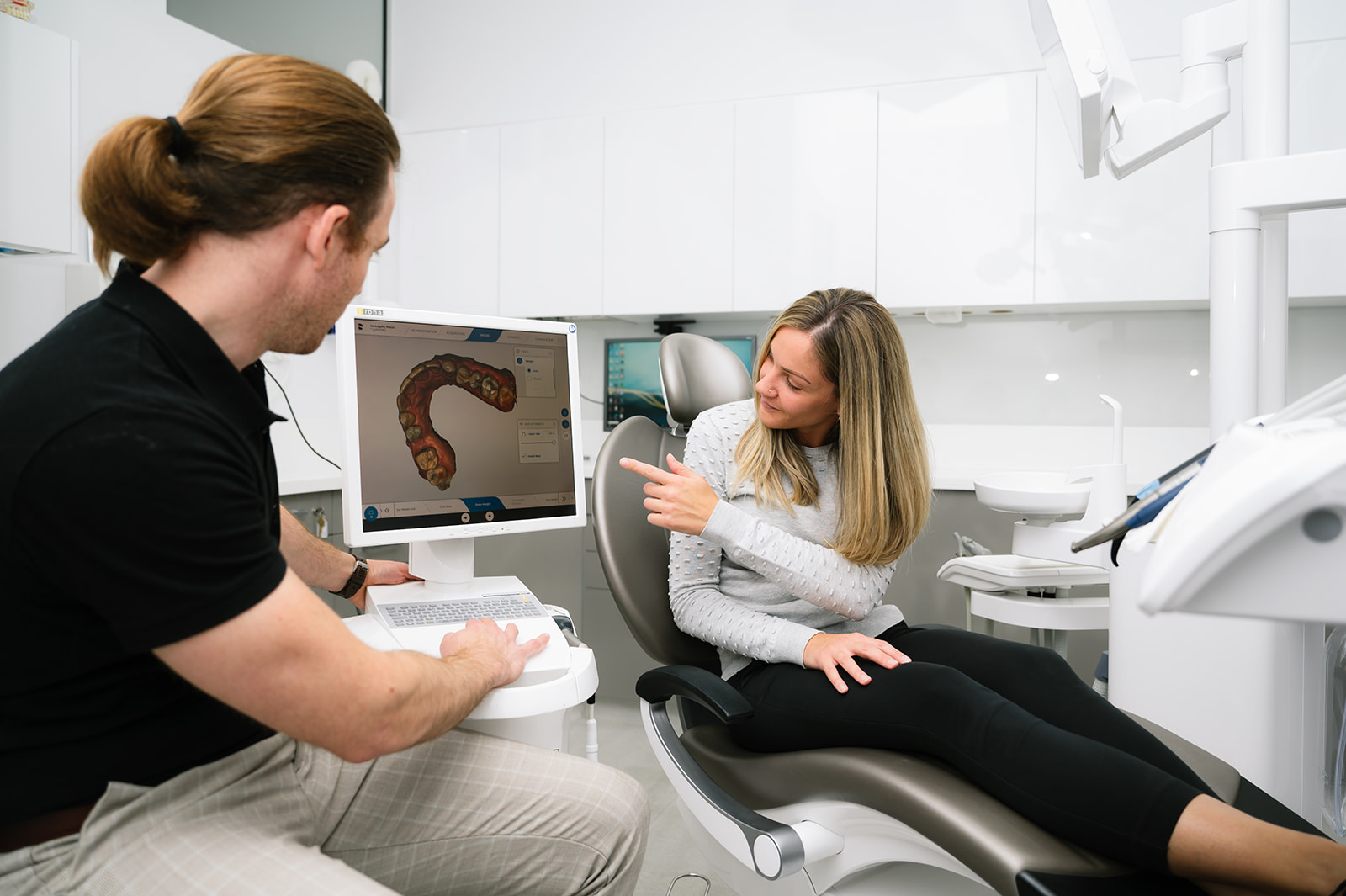 Male dentist showing a digital scan in a computer with the female patient sitting at the patient chair pointing at the computer