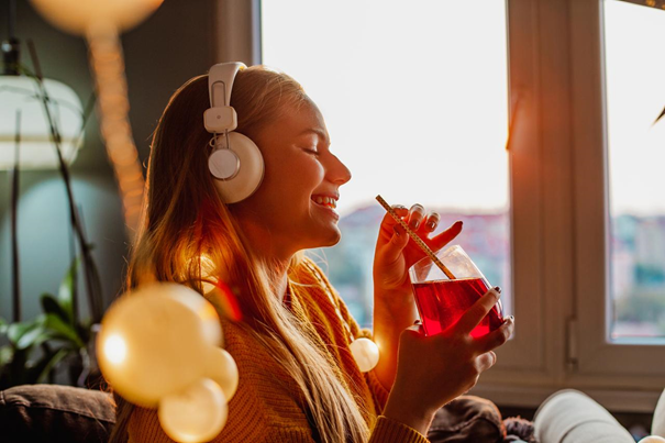 Eine junge Frau mit langen blonden Haaren sitzt in einem Loungesessel. Sie trägt einen gelben Strickpullover und weiße Kopfhörer. Sie trinkt mit Strohhalm ein rotes Getränk aus einem Cocktailglas.