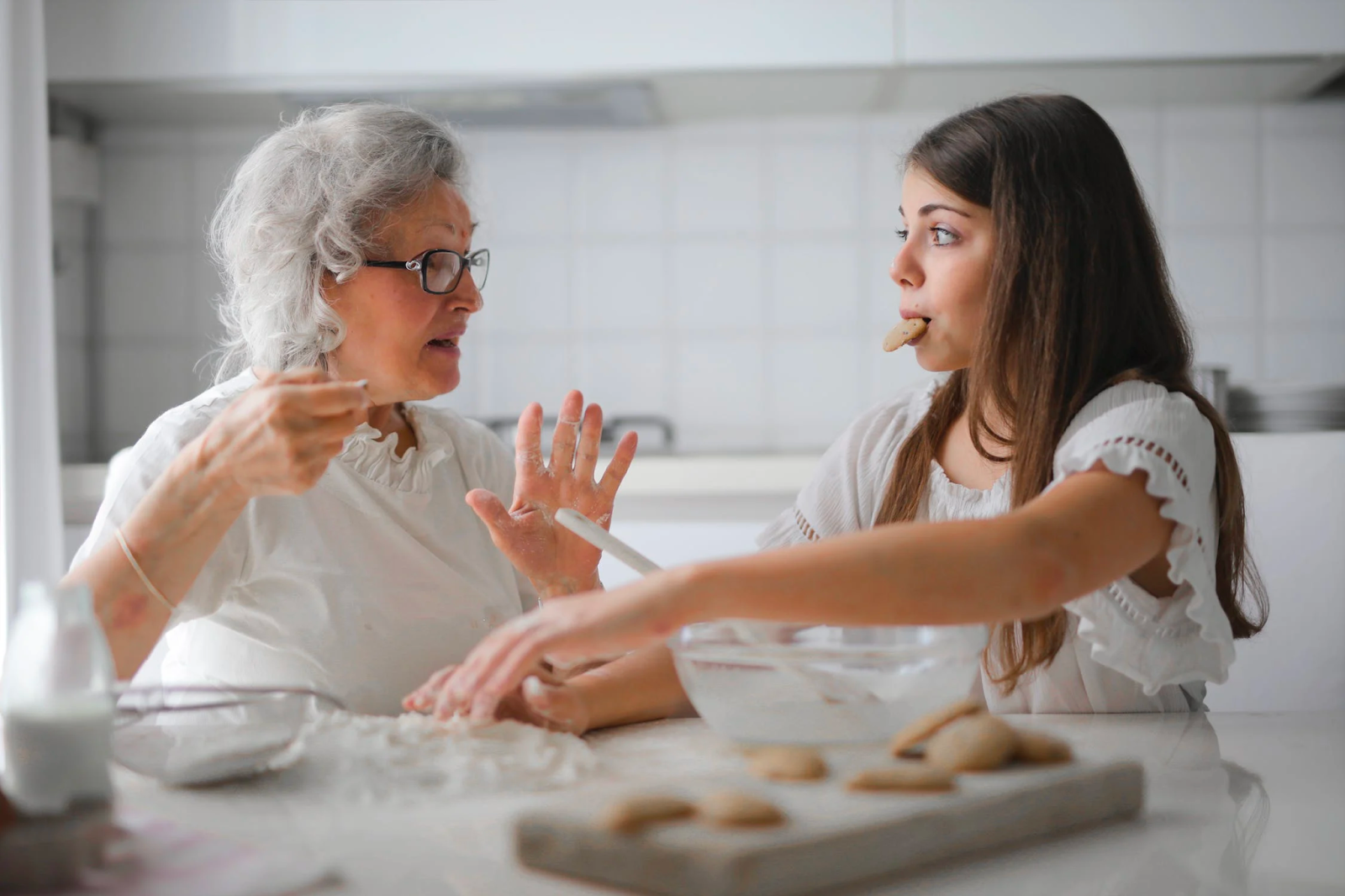 A grandmother teaching her granddaughter to bake. The granddaughter has a cookie in her mouth