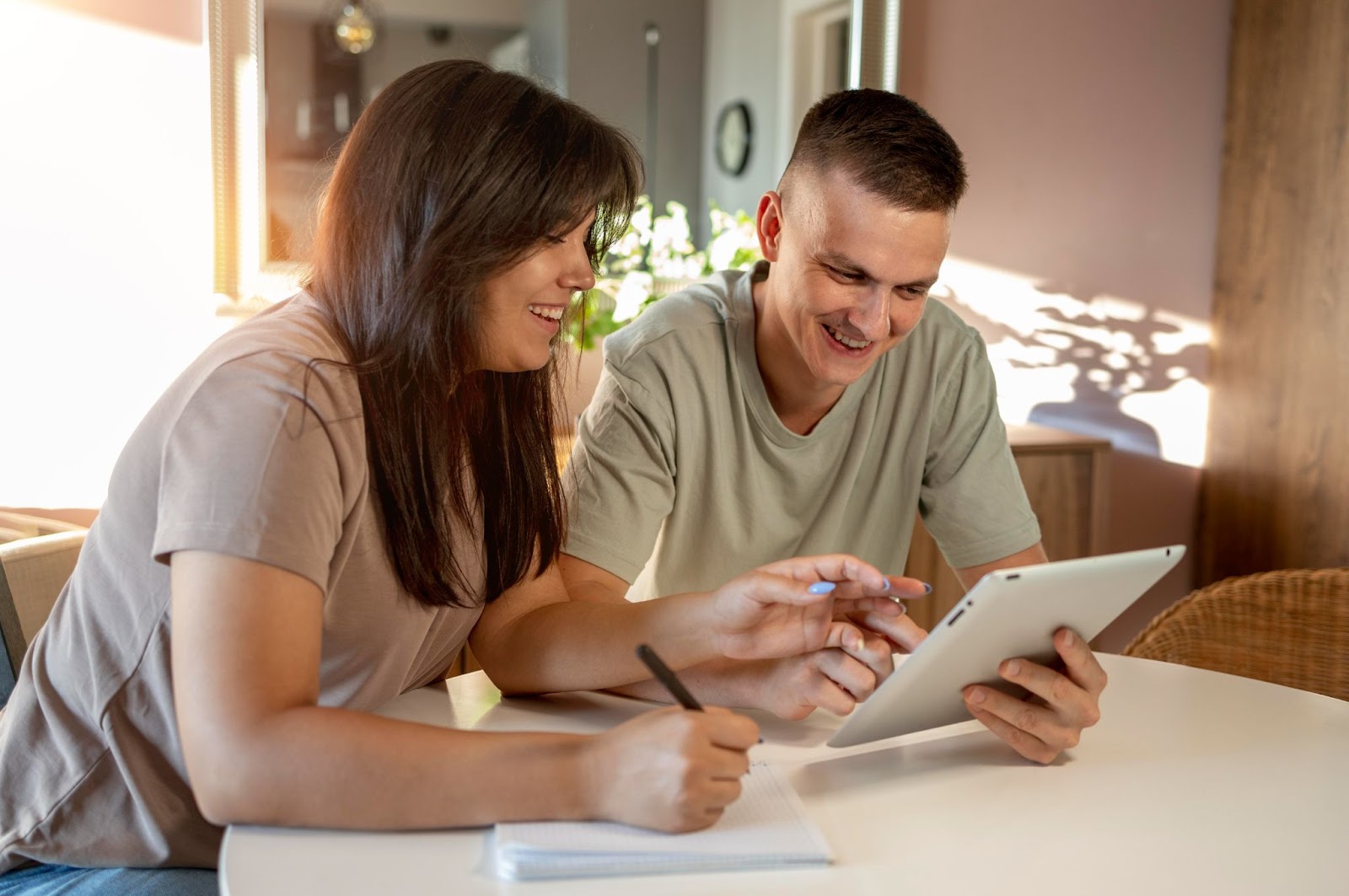 Imagem de um casal contente enquanto observa a tela de um tablet. A mulher está usando um caderno e uma caneta e o homem segura o eletrônico. Ambos estão sentados à mesa.