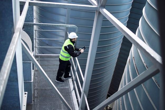 Factory silos worker standing on metal platform between industrial storage tanks and looking at tablet about food production