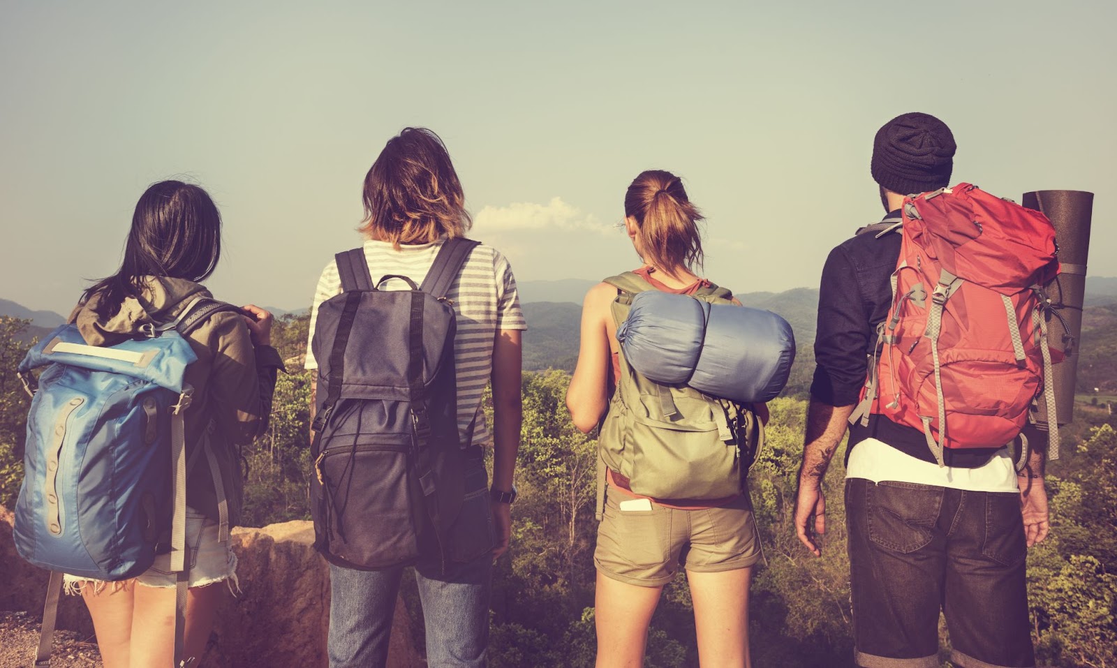 Grupo de viajantes observando uma paisagem serrana, composta por uma floresta verde e montanhas. As quatro pessoas estão de costas para a câmera e carregam mochilas de trilha.
