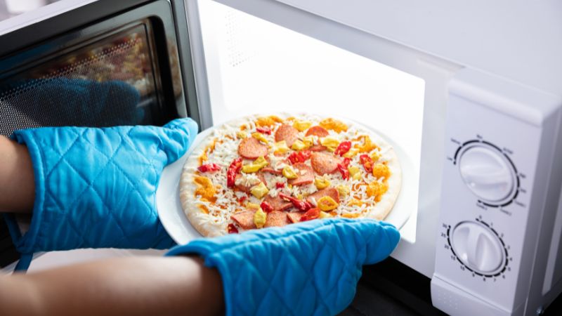 A person's hands wearing blue oven mitts are removing a white plate with an uncooked pizza topped with pepperoni, vegetables, and cheese from a microwave oven.