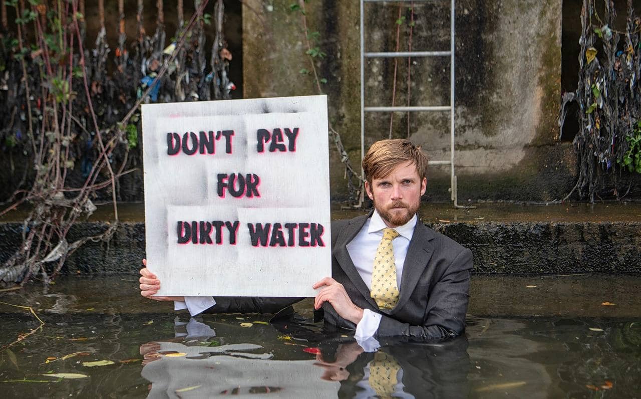 A rebel in a suit stands in a canal, water up to his torso, looking serious as he holds up a sign saying Don't Pay For Dirty Water