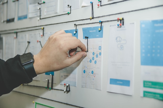 A person's hand pointing to a flowchart on a wall with other diagrams and documents pinned around it.