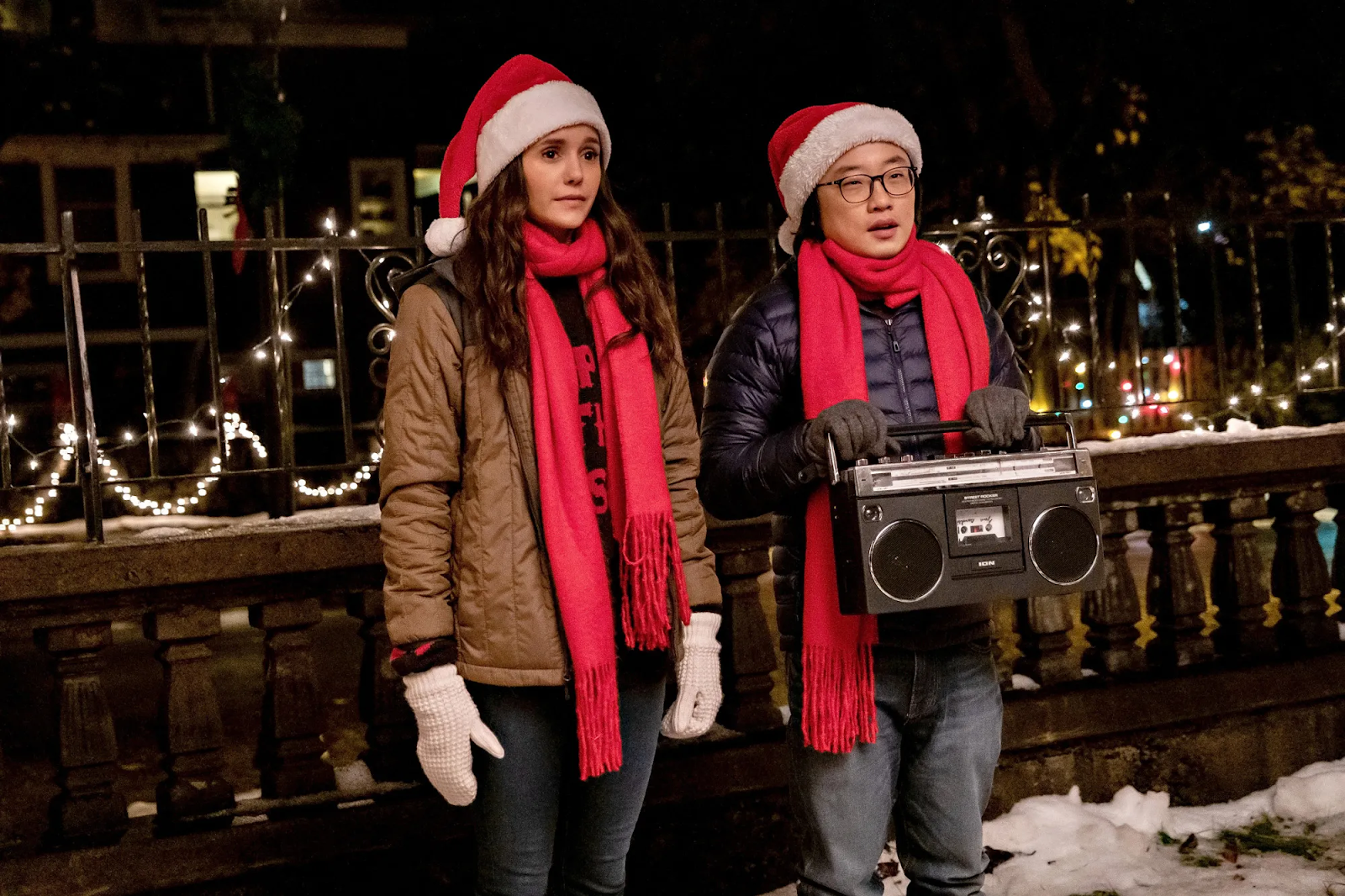 Jimmy O Yang and Nina Dobrev's characters in Love Hard stand outside, holding a boombox, sharing a heartfelt moment reminiscent of the tender and heartwarming scenes found in the best comfort Christmas films. Their faces show a mix of emotions, possibly conveying affection, vulnerability, or a romantic gesture, adding to the film's endearing charm.