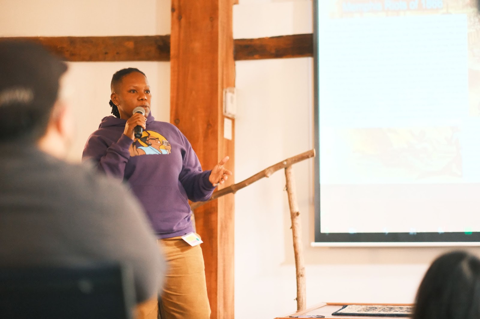 A black queer organizer LB wears a purple sweater with Ella Baker on it while speaking into a mic in front of a stage during their MJNF presentaiton