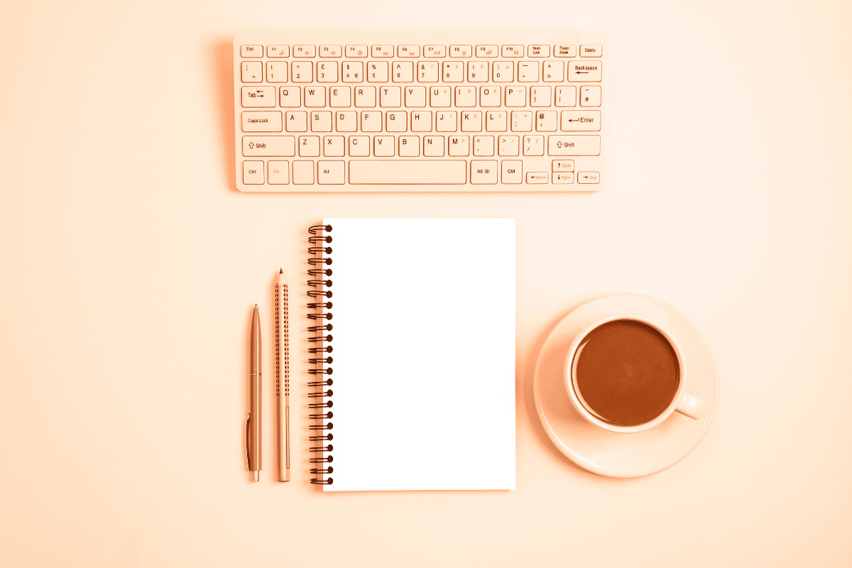 a flat lay scene of a keyboard, notepad, two pens, and a coffee mug on a peach fuzz colored backdrop