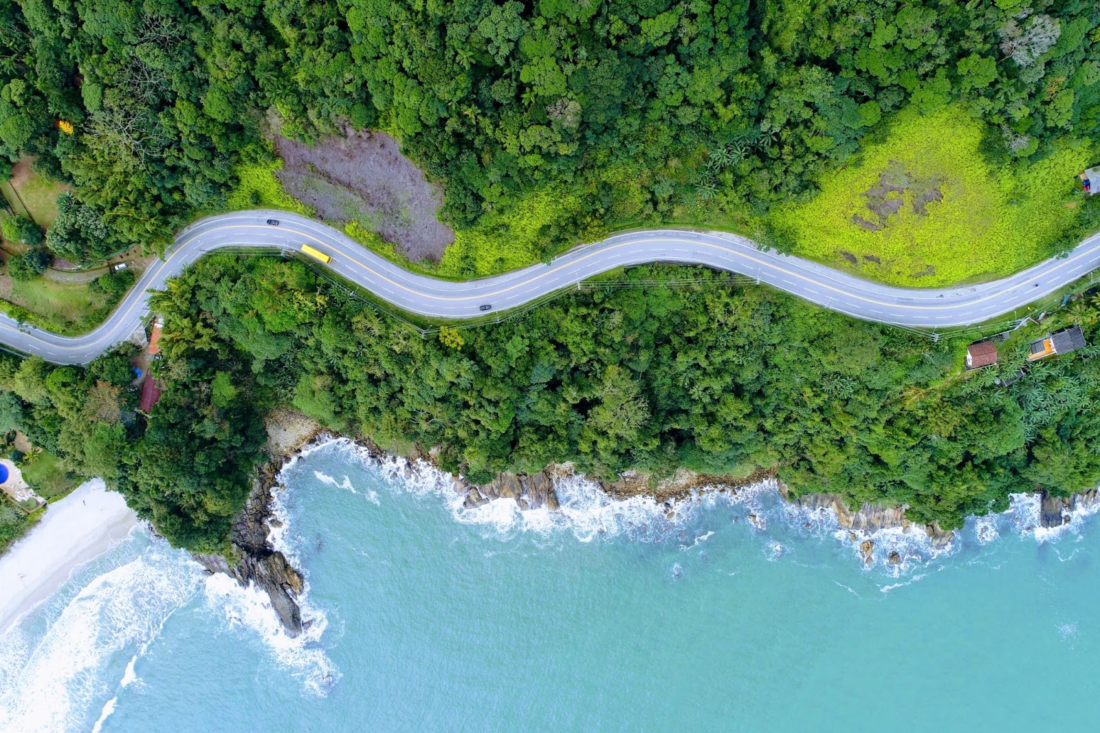 Trecho da Rodovia Rio-Santos, em São Sebastião. A estrada sinuosa passa por uma encosta densamente arborizada que é limitada pelo mar azul.