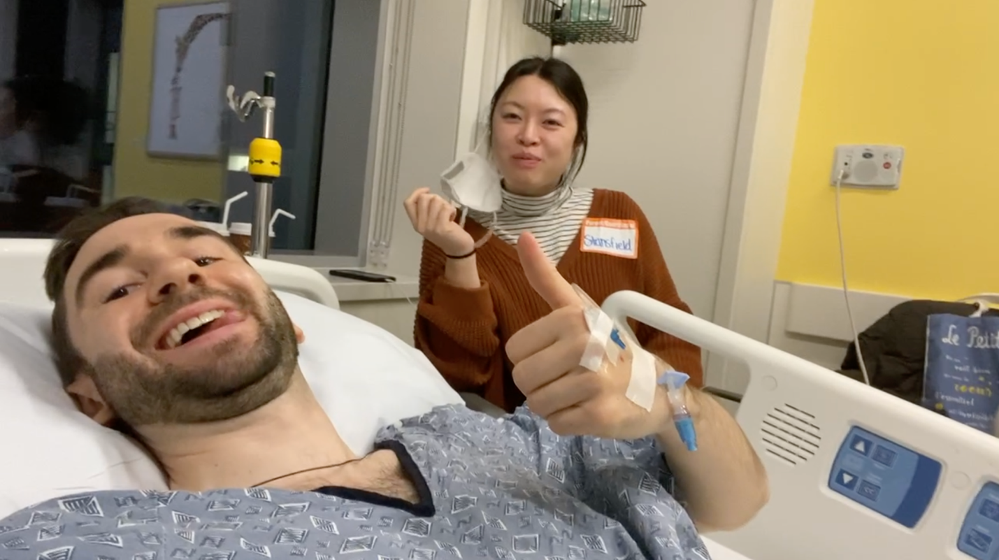 Stephan, lying on a hospital bed, gives a thumbs up. His wife smiles from a seat behind him.