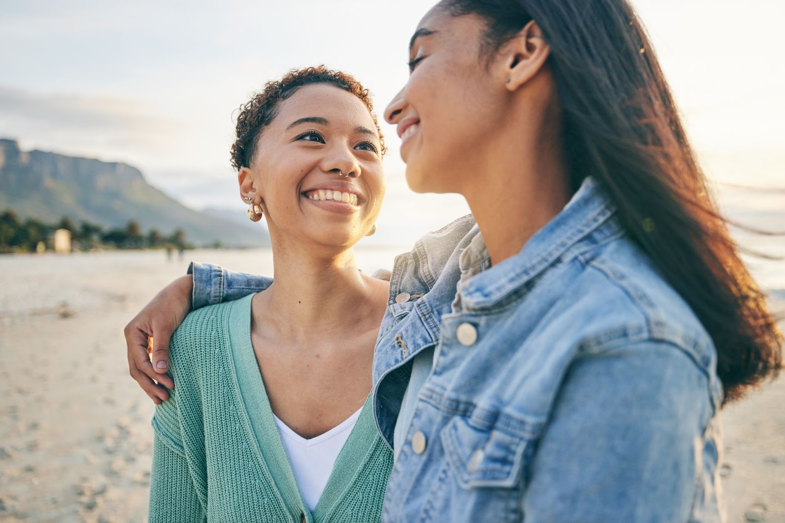 Casal composto por duas mulheres jovens caminhando pela areia de uma praia. Elas se abraçam lateralmente e sorriem uma para a outra.