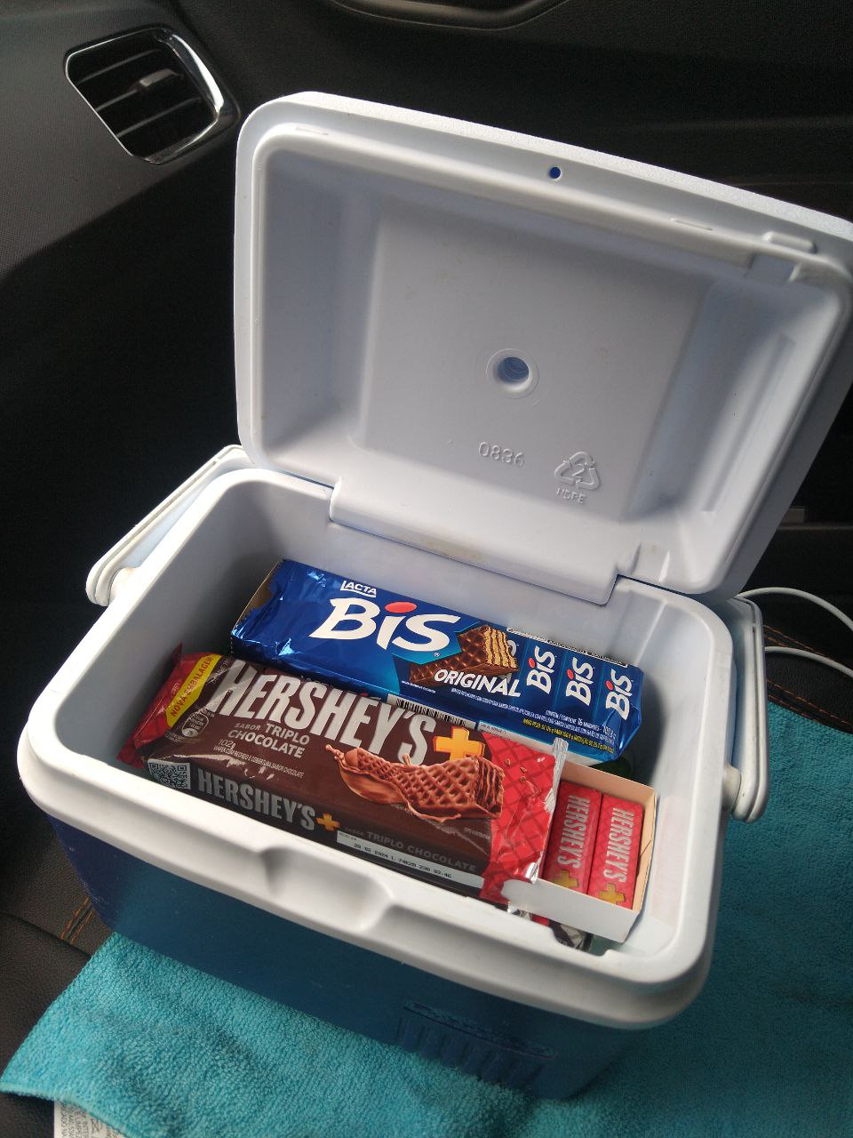 A imagem mostra o interior de um veículo, focado em um cooler branco pequeno aberto que está sobre o assento. Dentro do cooler, há várias barras de chocolate, incluindo marcas conhecidas como Hershey's e Bis. O cooler parece estar colocado sobre um assento com estofamento azul, sugerindo que pode ser uma comodidade oferecida aos passageiros, talvez em um serviço de transporte privado ou táxi.





