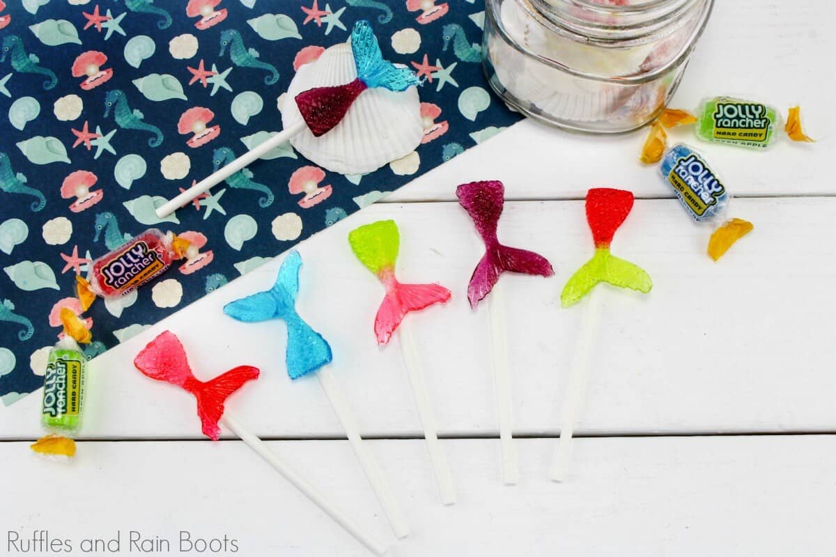 mermaid tail lollipops in multiple colors on a white background with some colorful paper and a shell with jolly rancher candies