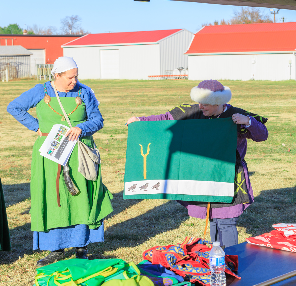 One woman showing off a green saddle pad with a gold pitchfork, a white band and three black ravens. A second woman observing.