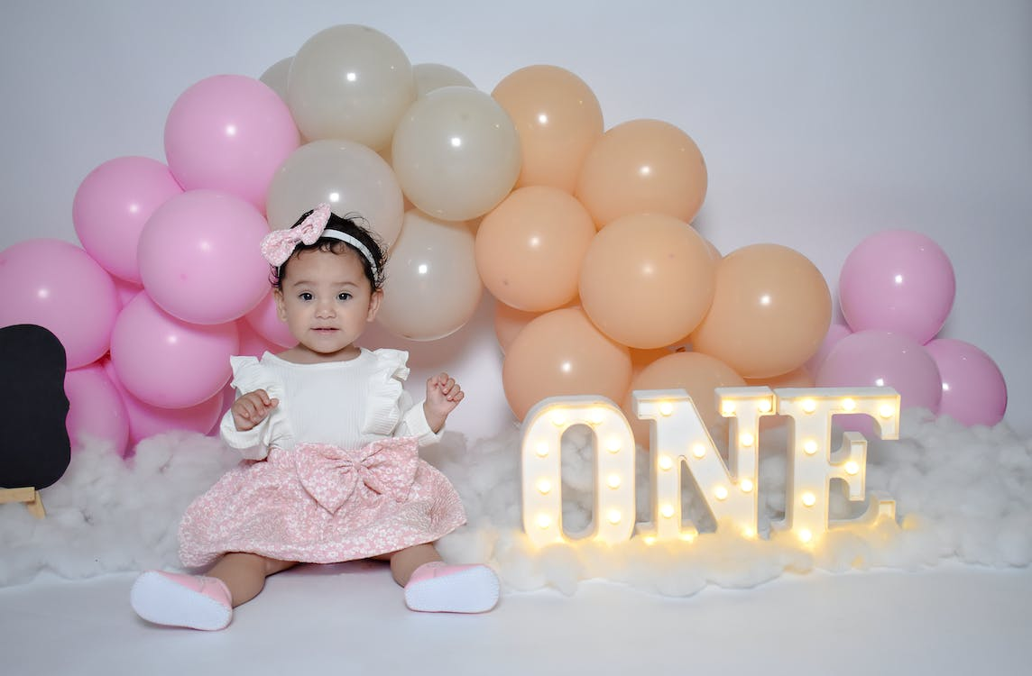 One year old adorable little toddler in a beautiful white and pick dress sitting in front of decorative balloons and beside 3D marquee lamps stating ONE. 