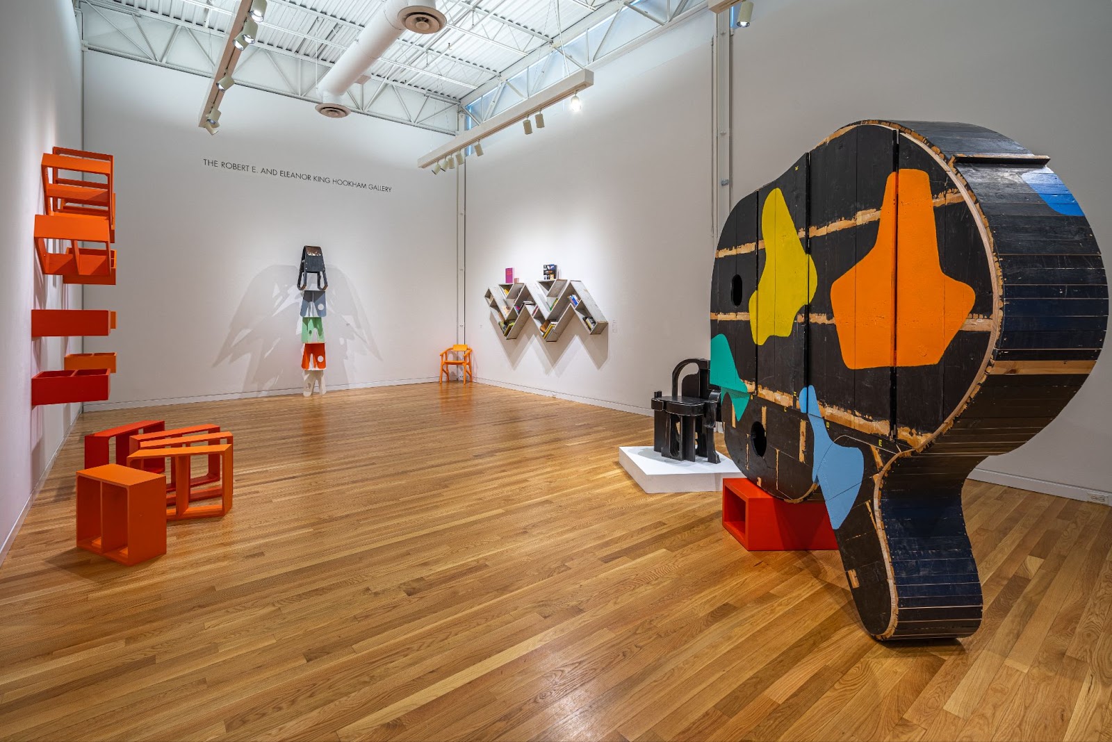 Image: Installation view of A Love Supreme. On the left side of the image, orange wooden boxes are placed on the ground and attached to the wall. On the right side, a large black wooden object is painted with colorful splotches. Two more pieces are mounted on the back walls of the exhibition space. Photograph by Siegfried Mueller, courtesy of the Elmhurst Art Museum. 