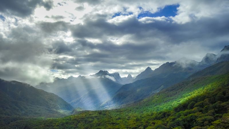 Sunbeams pierce through moody clouds over the rugged peaks and lush valleys of Fiordland National Park.