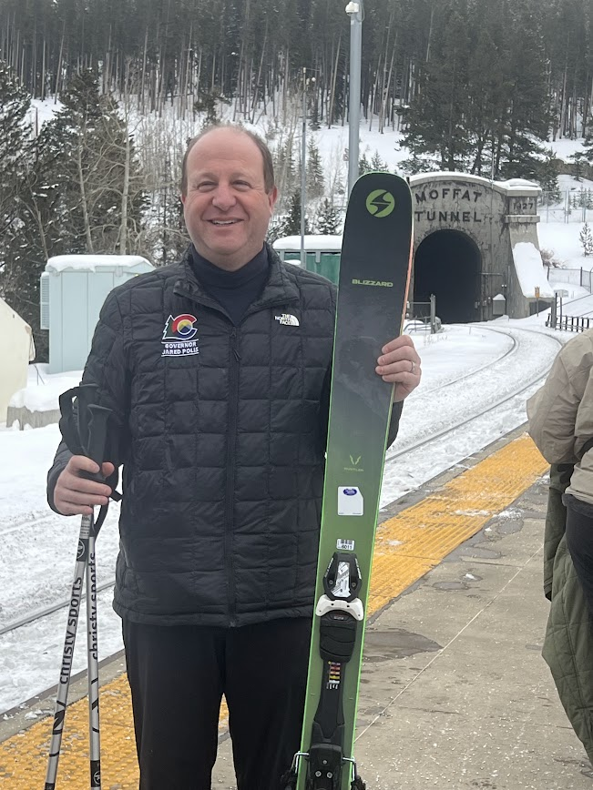 Governor Polis holding poles and skis with train tunnel in background.