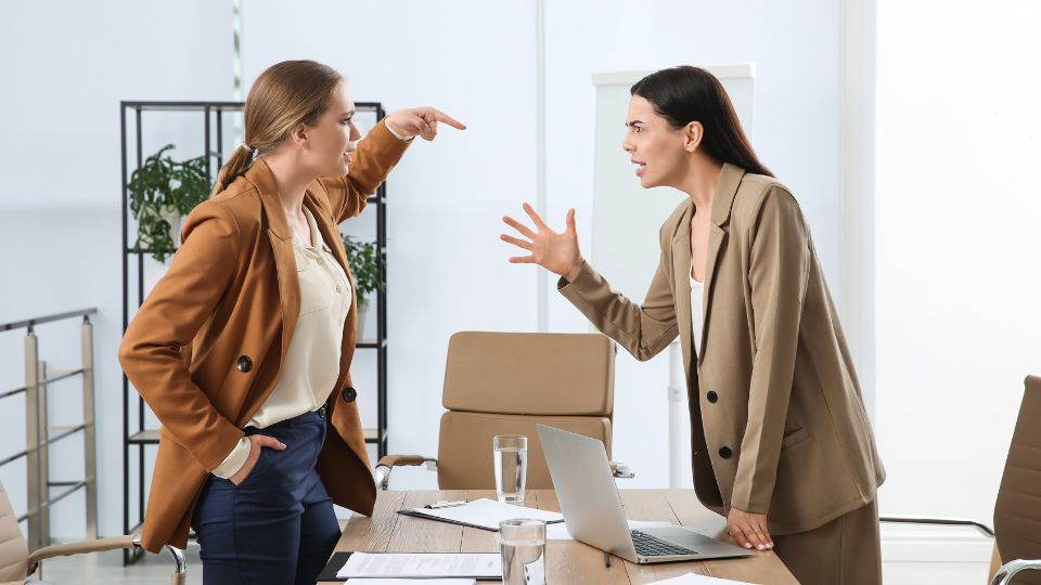 Two professional women in a modern office setting engaged in a heated discussion, with one pointing assertively and the other expressing a defensive stance.