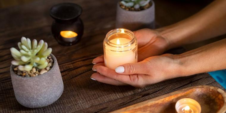 Close-up das mãos de uma mulher segurando uma vela aromática sobre uma mesa de madeira, rodeada por outras pequenas velas, enquanto relaxa em seu spa caseiro.