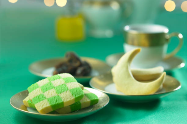 A festive arrangement of Hari Raya sweets with green layered cake, dates on a plate, and traditional cookies, accompanied by a cup of tea and a teapot in the background on a green tablecloth.