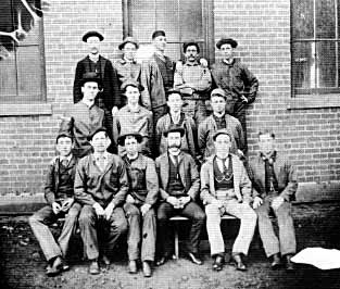 Group photo of fifteen men; five in the top row, four in the middle, and six in the front, posing in front of a brick building