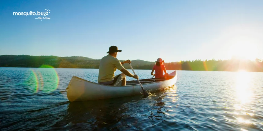 Man wearing a hat and a woman are canoeing on a lake as the sun either rises or sets.