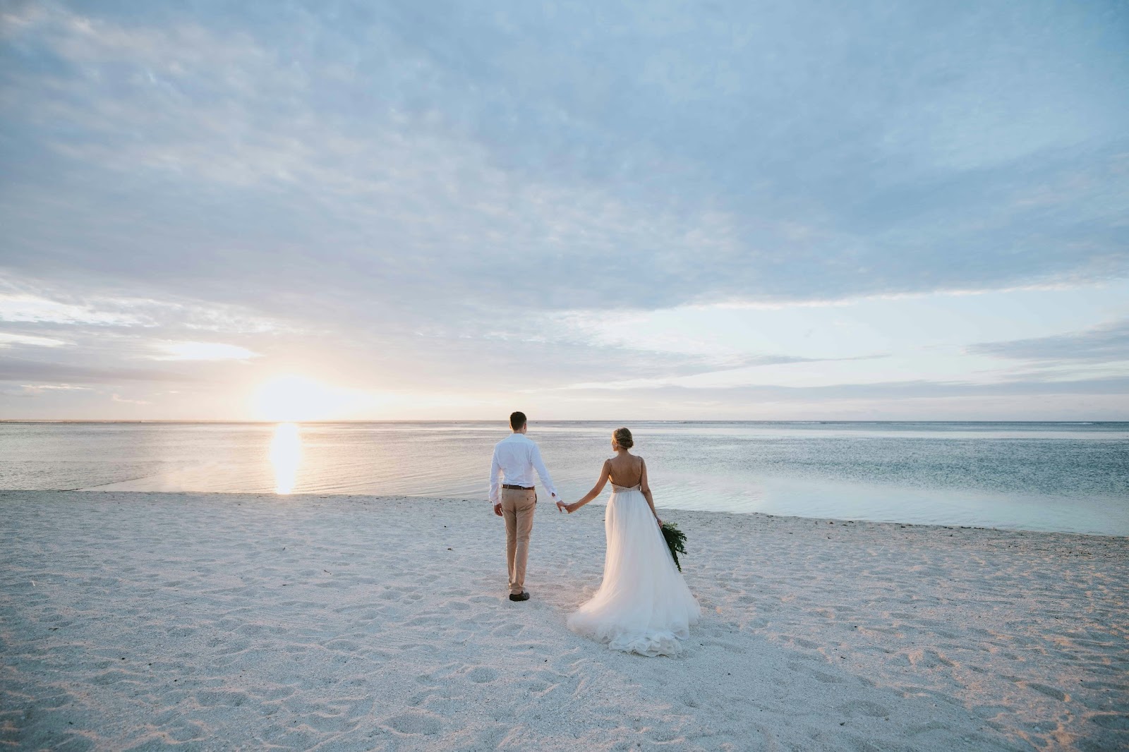 A bride and groom walking on the beach at dusk with clear blue skies and turquoise waters in the background.