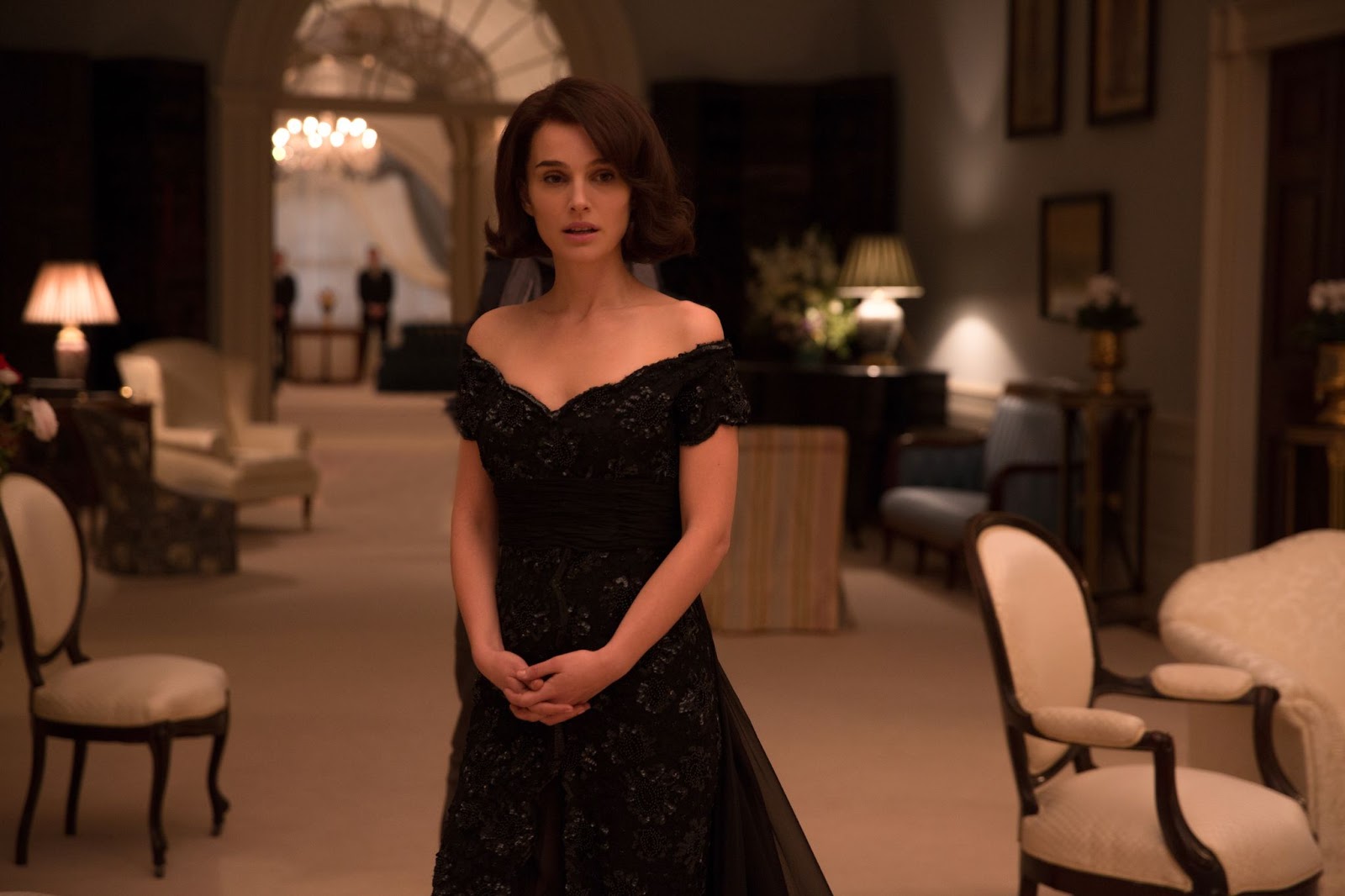 Natalie Portman como Jackie Kennedy, utilizando un vestido negro con los hombros descubiertos. Ella está de pie dentro de un salón