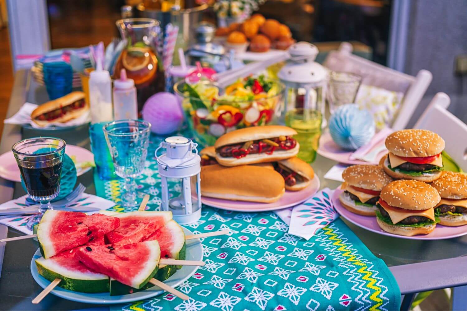 a buffet of burgers, hot dogs, and other pool party snacks on an outdoor patio
