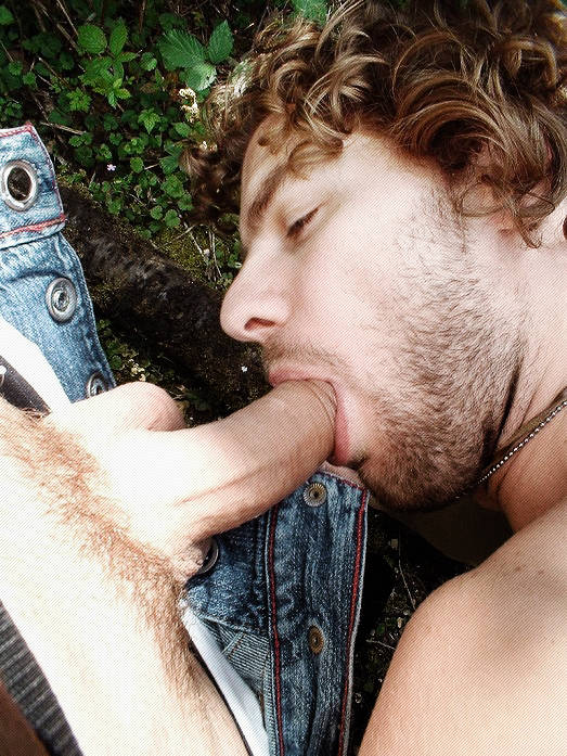 curly haired gay male sucking on a fat cut cock pulled out of the buttom fly of his boyfriend's jeans while hanging out outside
