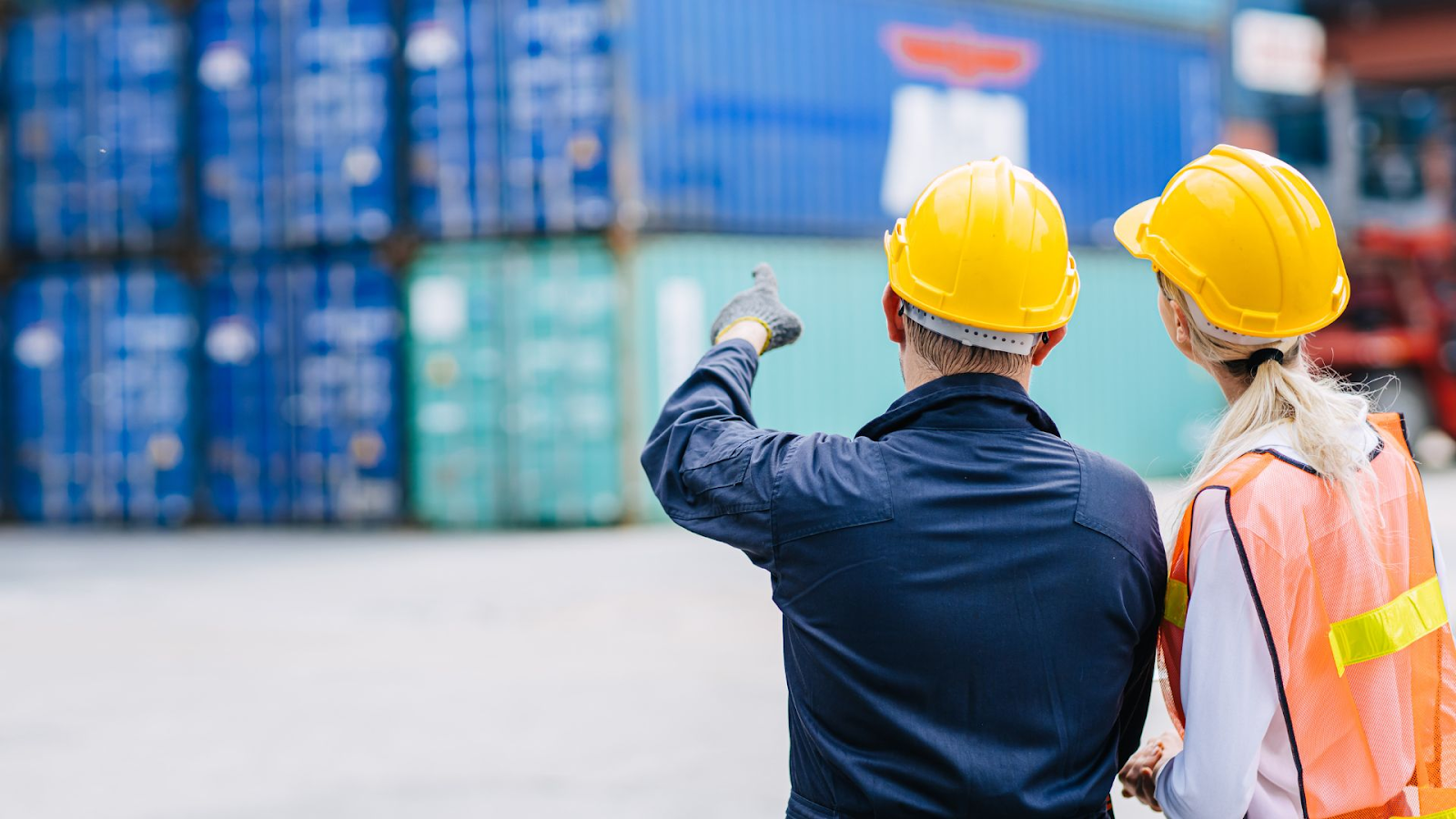 Rear view of two professionals at a shipping container yard. The person on the left, wearing a dark blue work uniform and a yellow safety helmet, is pointing towards something in the distance. The person on the right, wearing a reflective orange vest and a yellow safety helmet, is looking in the same direction. They are both standing in front of a backdrop of stacked blue shipping containers, indicative of an industrial port or logistics hub.