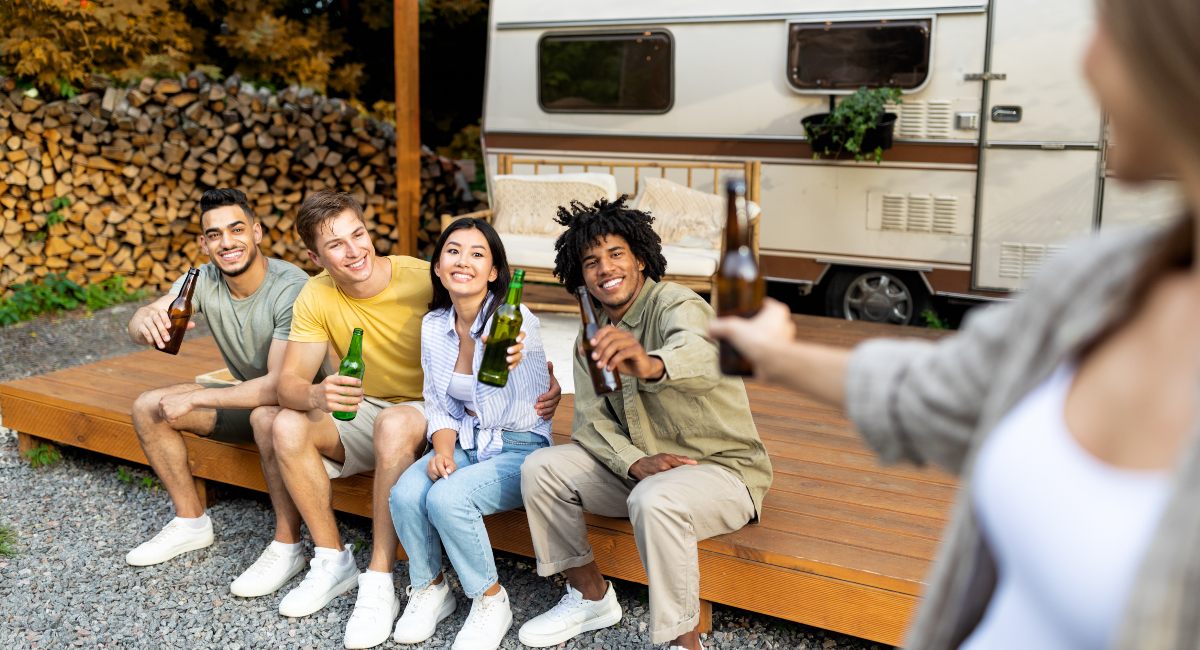 A group of cheerful multiracial friends toasting with beer bottles near a vintage RV in a camping setting, with a backdrop of firewood stacked and autumn trees, sharing a joyful outdoor gathering