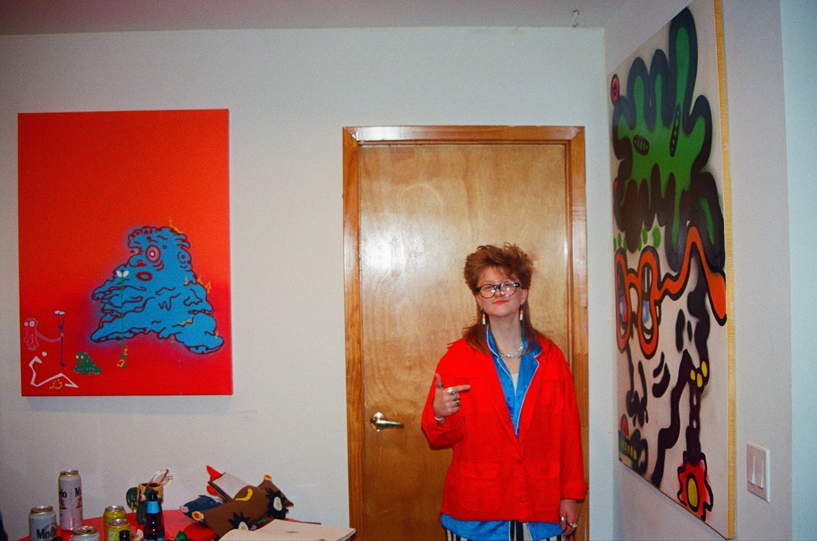 Alt text: Blonde girl wearing circular black glasses and a red jacket stands in front of a wooden door with a red and blue painting on the left and a white and green painting on the right.