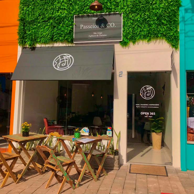 Fachada da agência de turismo "Passeios & CO.", com mesas e cadeiras de madeira na calçada e paredes decoradas com plantas.