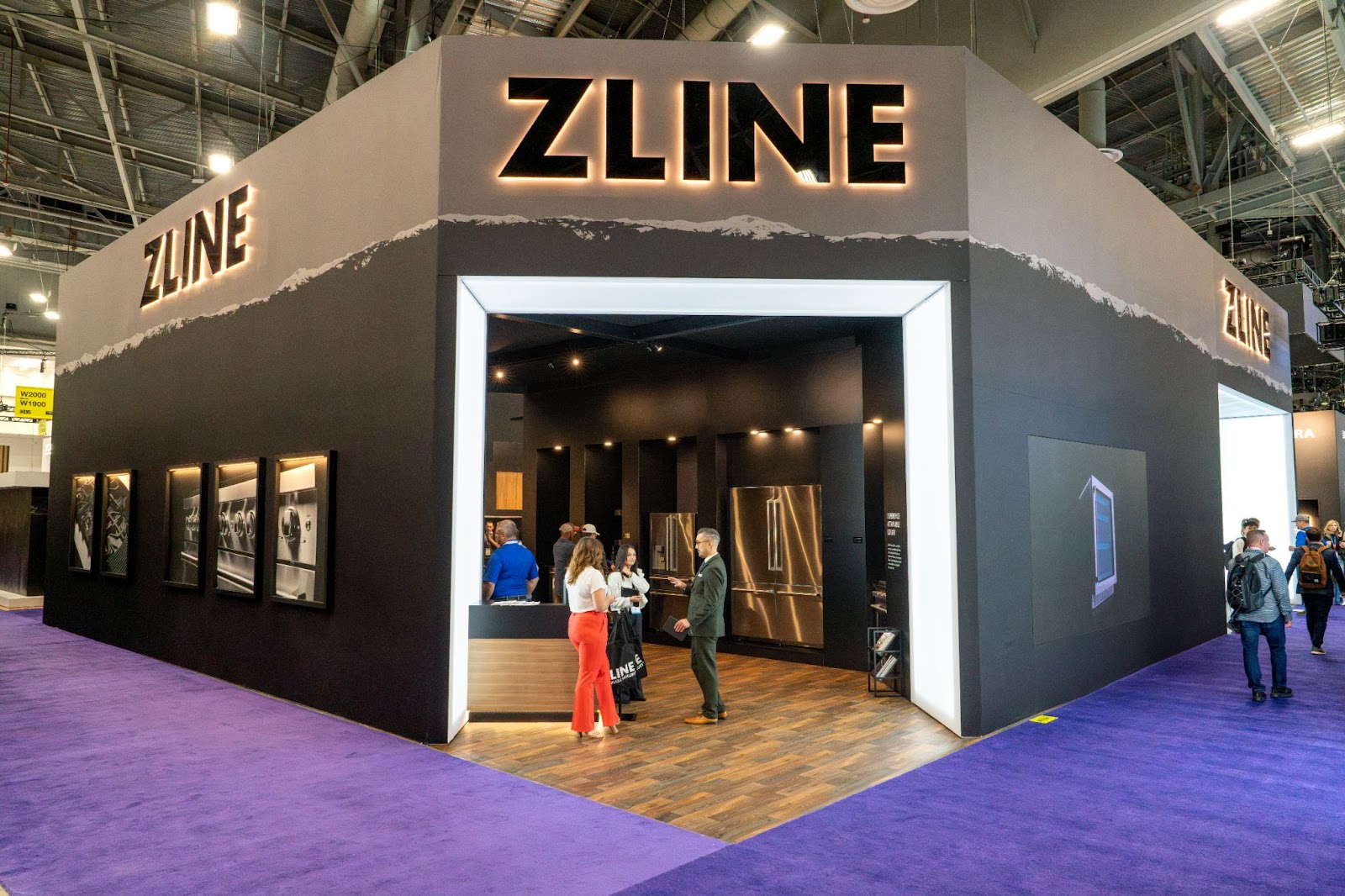 A trade show booth with prominent 'ZLINE Kitchen and Bath' branding on its facade, featuring visitors and staff. The booth is designed with a dark exterior, illuminated company logo, and a welcoming entrance framed by mountain peak graphics.