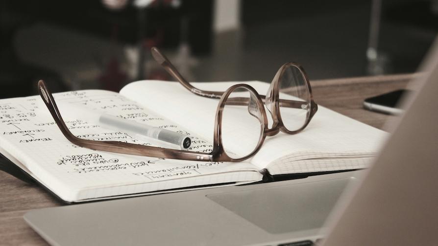 A pair of eyeglasses beside a pen, both on top of a notebook with scribble while a laptop is also open -- tools used by an organizer looking for female inspirational speakers.