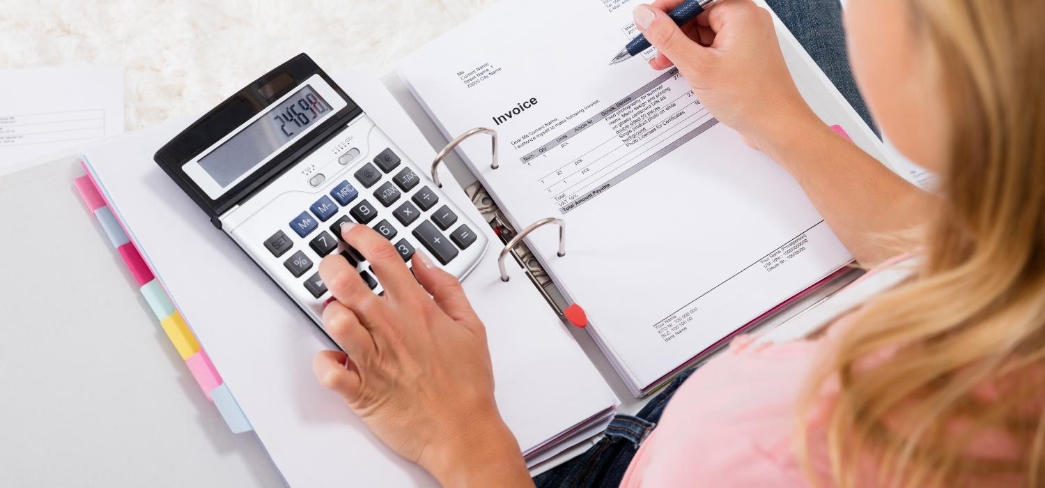 Seorang wanita pengusaha yang mengenakan kemeja merah muda dengan rambut pirang sedang menghitung biaya untuk anggaran perjalanan bisnis dari perusahaan GOTAP menggunakan kalkulator.